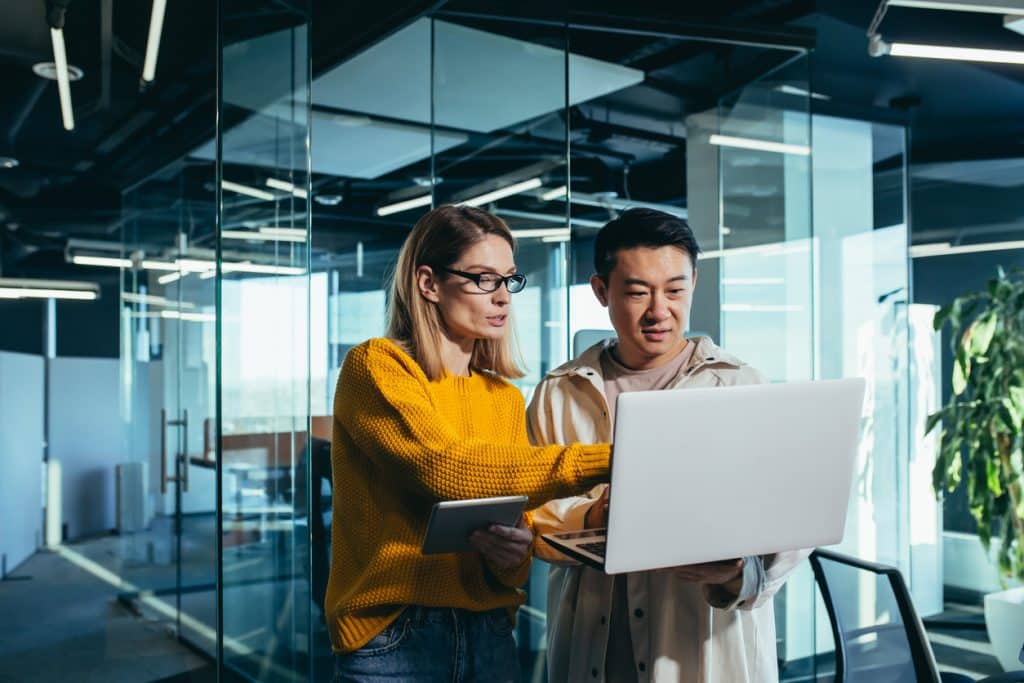 Business meeting of two employees, Asian and female colleagues, discussing the project, working in a modern office, looking at a laptop screen, discussing edits, and project success